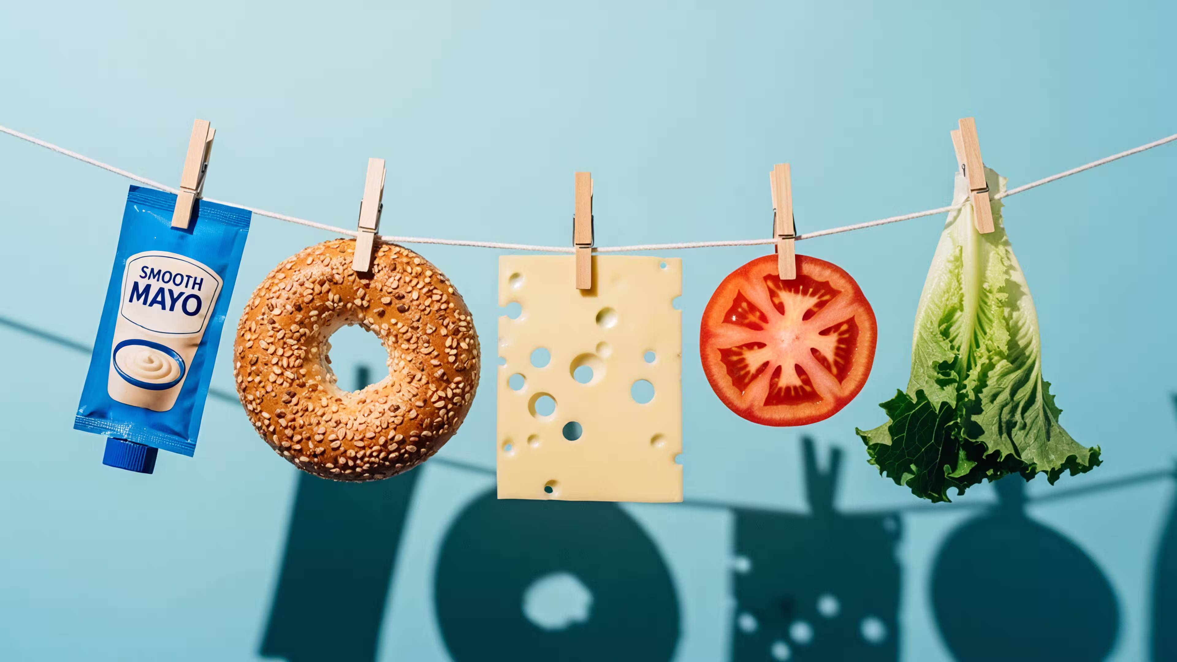 Ingredients for a sandwich clipped to a clothesline: mayo, bagel, cheese, tomato, and lettuce.
